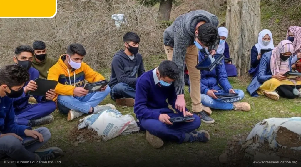 A group of students in Kashmir are studying in an outdoor class on tablets from Tab Lab. They are sitting on a mountainside under the shade of a tree